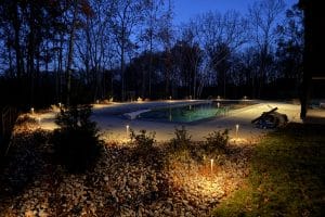 IMG_6912 | LandCo Group Nighttime backyard scene: illuminated swimming pool with warm pathway lights along a rocky border and leafless trees in the background.