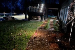 IMG_6913 | LandCo Group Nighttime backyard with a raised wooden deck, a mulched flower bed edged by small shrubs, illuminated path lights, and a pool visible on the left.