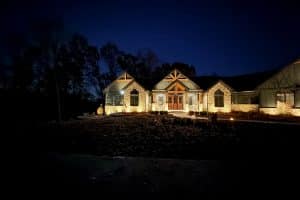IMG_6919 | LandCo Group Two- story stone house at night with warm exterior lights along the entrance and front path, trees in the dark background, and a clear night sky.
