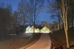 IMG_6922 | LandCo Group Illuminated house at night at the end of a curved path, with bare trees and a light dusting of snow along the ground.