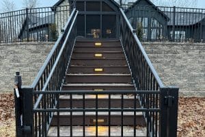 IMG_7021 | LandCo Group Outdoor metal staircase with illuminated steps leading to a dark-framed building, brick retaining wall and gate at the bottom.