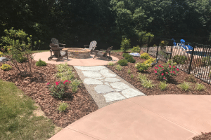 Outdoor patio with a circular stone fire pit surrounded by wooden Adirondack chairs on a brick patio, connected by a stone path through mulched flower beds.