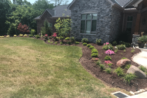 Front yard with a curved mulch flower bed, green shrubs and pink blooms along a stone-front house.