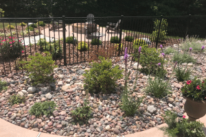 Rock garden with shrubs and purple blooms, separated by a black metal fence from a circular stone patio with a fire pit and chairs; a pot of red flowers sits on the right.