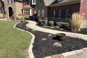 Curved concrete walkway through mulched landscaping in front of a brick-and-stone house, with trimmed shrubs and ornamental grasses.