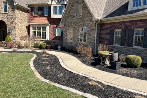 Curved concrete walkway bordered by stone edging winds through a landscaped front yard of brick-and-stone houses and trimmed shrubs.