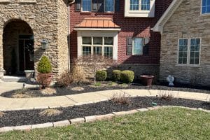 Front yard of a brick-and-stone house: arched stone entry, windows, shrubs, mulch beds, and a curved concrete walkway.