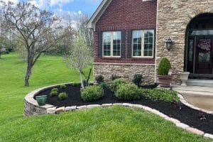 Curved mulch garden bed with small shrubs and a young tree in front of a brick-and-stone house, green lawn to the left on a sunny day.