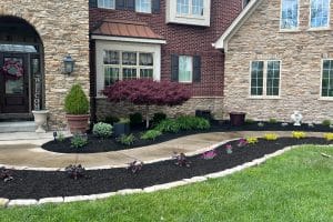 Front yard landscape with curved concrete path, mulched flower beds, and a red Japanese maple in front of a brick-and-stone house.