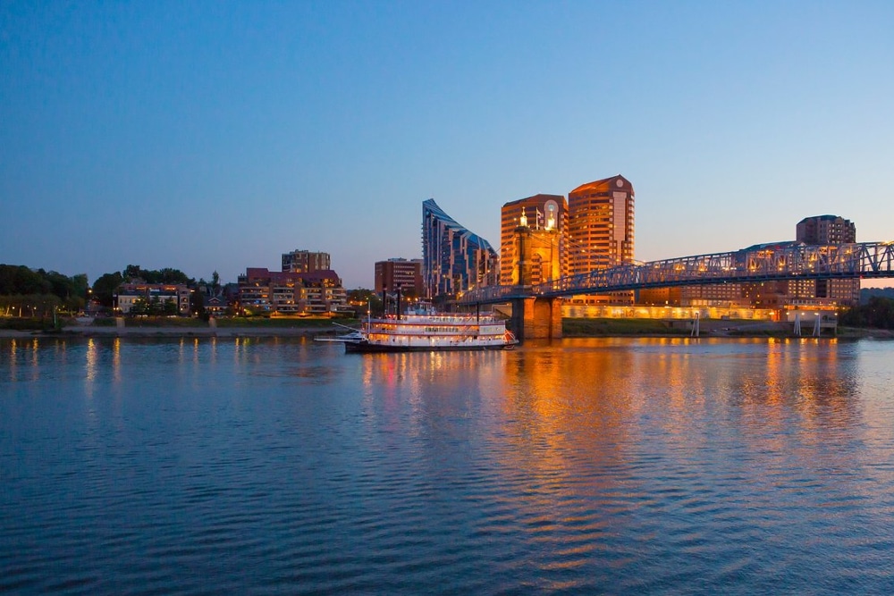 Covington-Kentucky | LandCo Group Twilight city skyline along a calm river, with a blue steel bridge and a lit boat on the water.