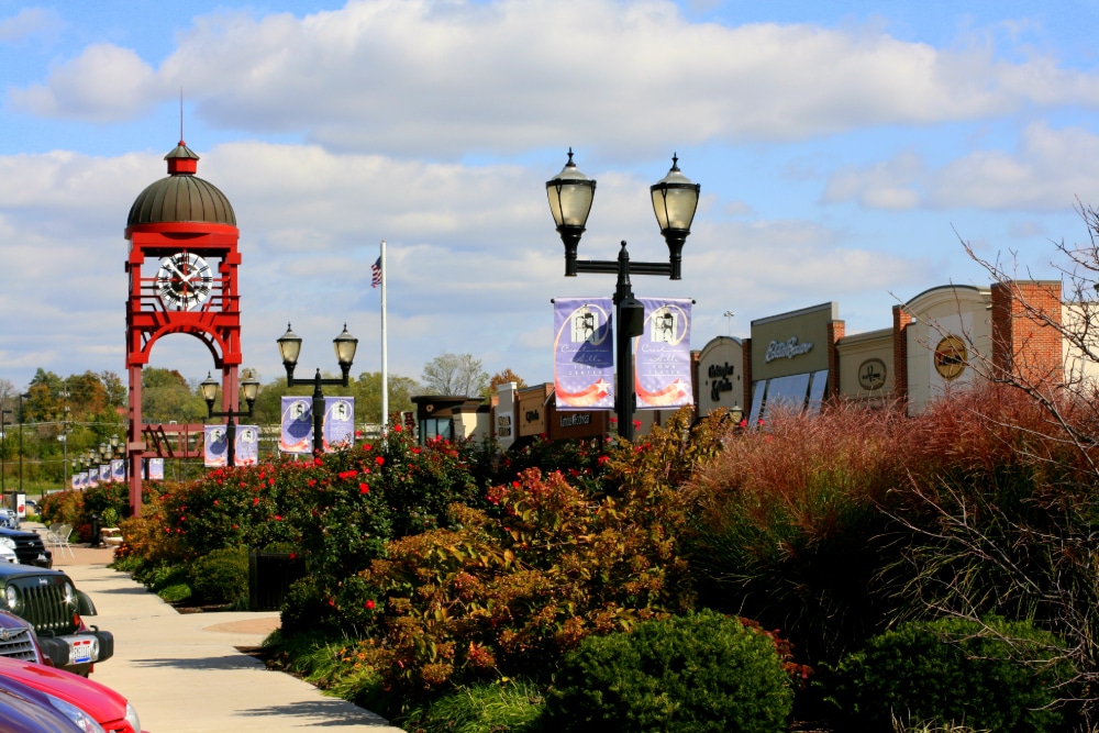 Crestview-Hills-Kentucky | LandCo Group Red clock tower structure along a shopping street with banners and lampposts under a blue sky with clouds.