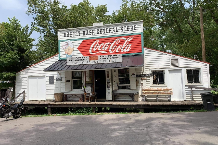 Rabbit Hash | LandCo Group White wooden general store with a large Coca-Cola sign and 'Rabbit Hash General Store' on the roof sign, set among trees with a porch and bench.