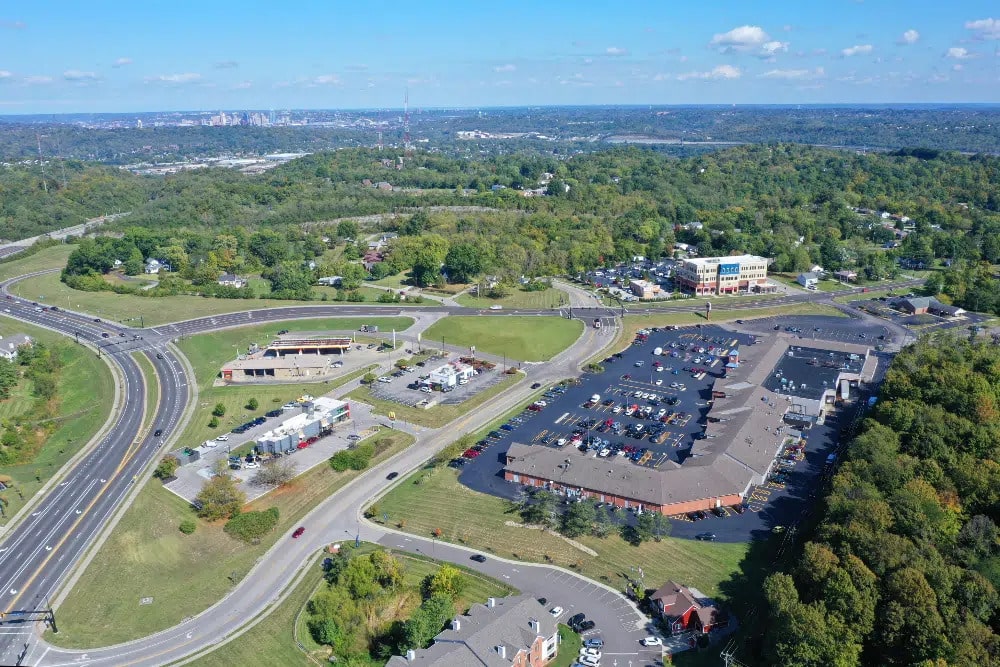 Taylor-Mill-Kentuckyjpg | LandCo Group Aerial view of a suburban commercial area with a large parking lot, a circular road interchange, and surrounding green trees.