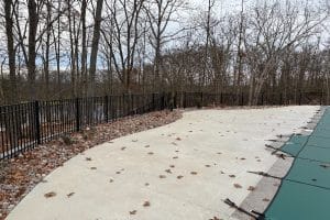 Union Full Landscape 10 | LandCo Group Empty concrete patio with fallen leaves, black metal fence, and a covered pool on the right; leafless trees in the background.