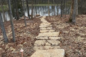 Union Full Landscape 13 | LandCo Group Stone stepping-stone path leading through fallen leaves toward a calm lake in a forest clearing by the water's edge