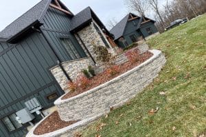 Union Full Landscape 5 | LandCo Group Curved stone retaining wall with mulch and small trees in front of dark gray modern houses, a leaf-covered lawn in the foreground.