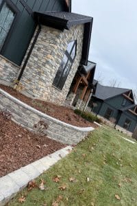 Union Full Landscape 7 | LandCo Group Modern house exterior with stone veneer, dark green siding and wood accents; a curved stone retaining wall with mulch beds and fallen leaves in the front yard.