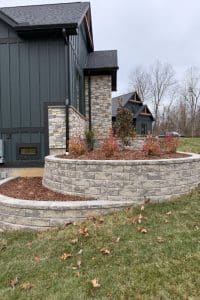 Union Full Landscape6 | LandCo Group Curved stone retaining wall with mulch bed and shrubs beside a dark-gray house with stone accents.